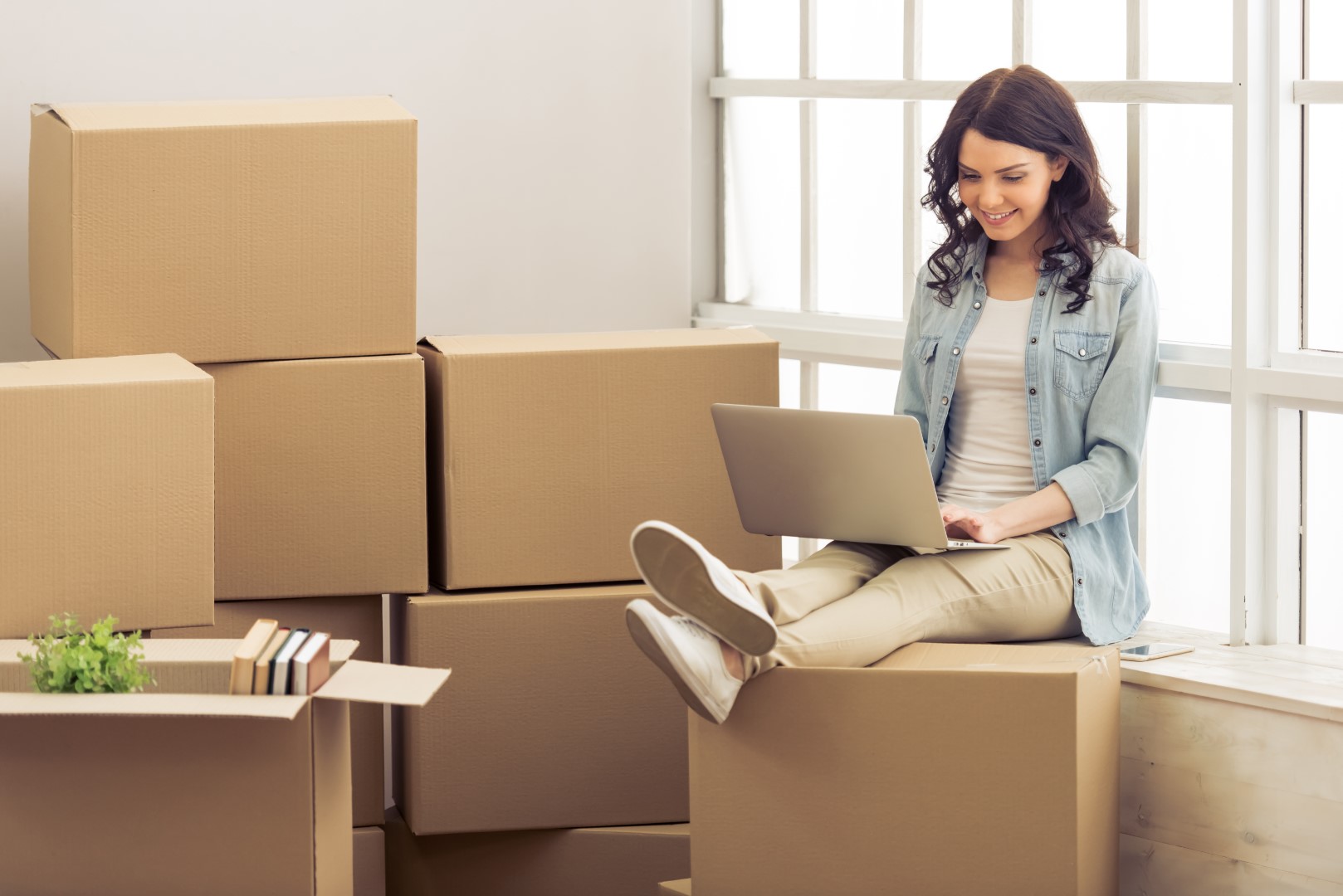 Young woman is moving, sitting among cardboard boxes, using a laptop and smiling