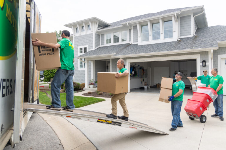 Movers in green shirts carry boxes and bins up a ramp into a moving truck outside a suburban house.